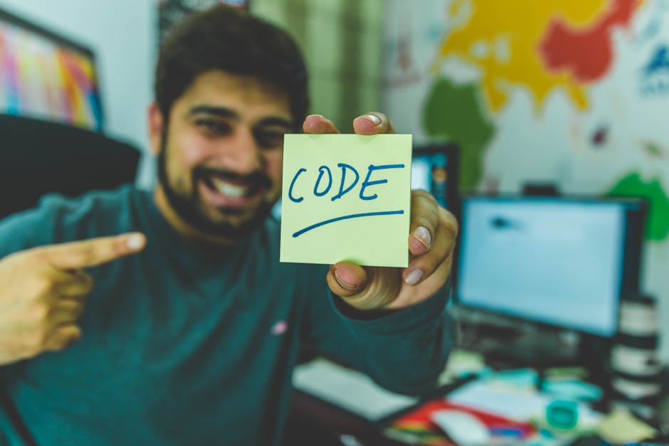 pexels-photo-879109-879109 A smiling programmer pointing to a sticky note with 'code' written on it in an office setting.